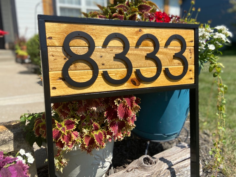 May include: Black metal house number sign with the number 8333 on a wooden plank. The sign is mounted on a black metal frame and is surrounded by plants.