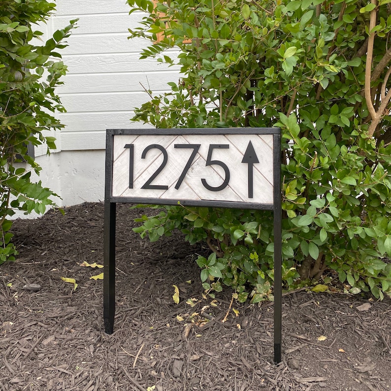 May include: A black metal address sign with the number "1275" and an upward-pointing arrow. The sign has a white and gray wooden background and a black frame. The sign is mounted on two black metal posts.