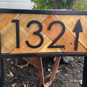 May include: Wooden address sign with the number 132 and an arrow pointing up. The numbers and arrow are black and mounted on a wooden background with a black metal frame. The wood has a natural, rustic appearance.