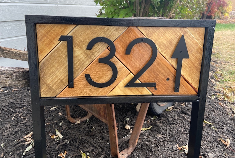May include: Wooden address sign with the number 132 and an arrow pointing up. The numbers and arrow are black and mounted on a wooden background with a black metal frame. The wood has a natural, rustic appearance.
