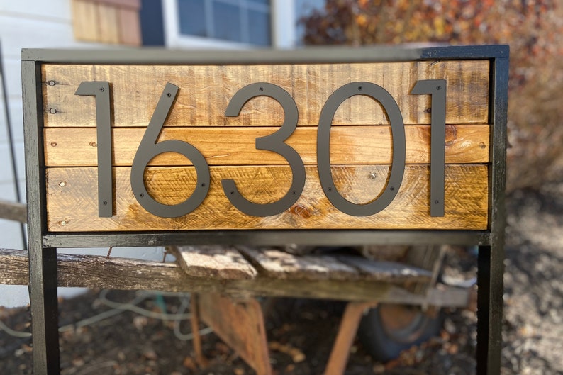 May include: A wooden sign with black metal house numbers reading "16301". The sign is mounted on a black metal frame and is standing on a wooden surface.
