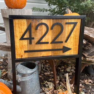 May include: Wooden address sign with the number "4227" in black, with an arrow pointing right. The sign is framed in black metal and mounted on a stand. The background includes pumpkins and a watering can.