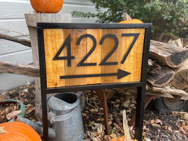 May include: Wooden address sign with the number "4227" in black, with an arrow pointing right. The sign is framed in black metal and mounted on a stand. The background includes pumpkins and a watering can.