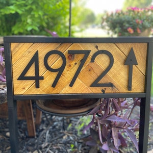 May include: A wooden address sign with the number 4972 and an arrow. The numbers and arrow are black and mounted on a light brown, chevron-patterned wooden background. The sign is framed in black metal.