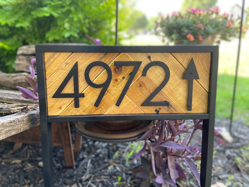 May include: A wooden address sign with the number 4972 and an arrow. The numbers and arrow are black and mounted on a light brown, chevron-patterned wooden background. The sign is framed in black metal.