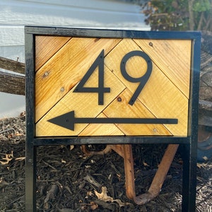 May include: A wooden address sign with the number 49 in black, with an arrow pointing left. The sign has a black metal frame and a chevron pattern made of light-colored wood. The sign is mounted on a black metal stand.