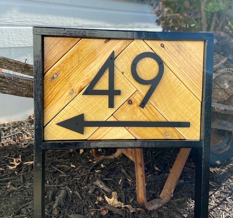 May include: A wooden address sign with the number 49 in black, with an arrow pointing left. The sign has a black metal frame and a chevron pattern made of light-colored wood. The sign is mounted on a black metal stand.