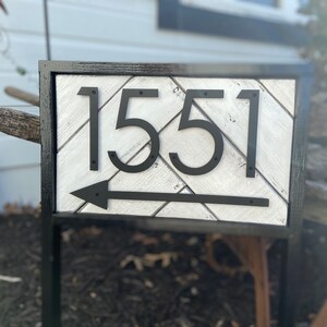 May include: A rectangular address sign with the number 1551 in black, stylized font. The sign has a white and gray chevron pattern background, a black arrow, and a black frame. The sign is mounted on a black metal stand.