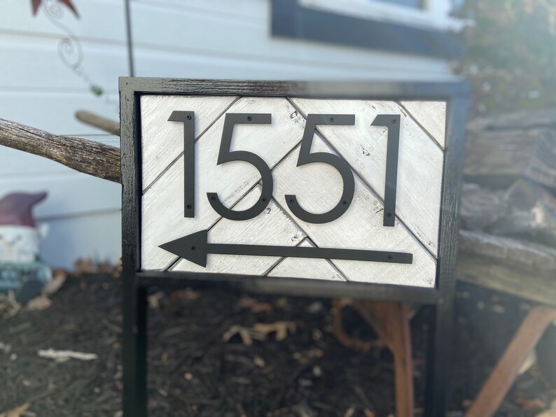 May include: A rectangular address sign with the number 1551 in black, stylized font. The sign has a white and gray chevron pattern background, a black arrow, and a black frame. The sign is mounted on a black metal stand.