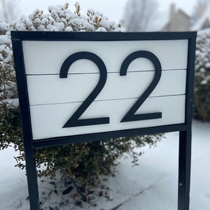 May include: A black metal frame with a white wooden sign displaying the number 22. The sign is mounted on a wooden post and is surrounded by snow-covered bushes.