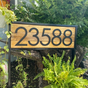 May include: Black metal house number sign with the number 2358 attached to a wooden plank. The sign is mounted on a black metal frame and is surrounded by green foliage.