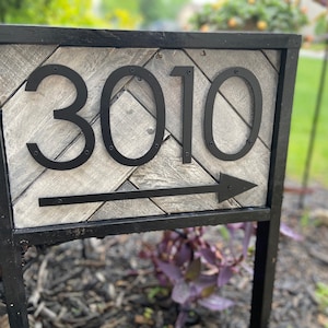 May include: A black metal address sign displaying the number 3010 with an arrow. The numbers are large and bold, mounted on a gray and white chevron-patterned wooden background. The sign is framed in black and mounted on a black metal post.