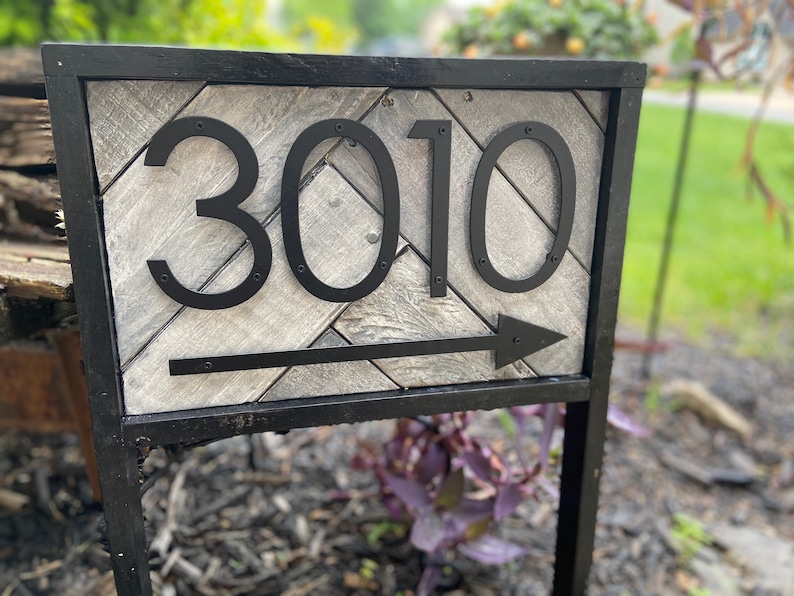 May include: A black metal address sign displaying the number 3010 with an arrow. The numbers are large and bold, mounted on a gray and white chevron-patterned wooden background. The sign is framed in black and mounted on a black metal post.