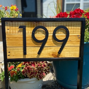 May include: Black metal house number sign with the number 199 on a wooden background. The sign is mounted on a black metal frame and is surrounded by flowers.