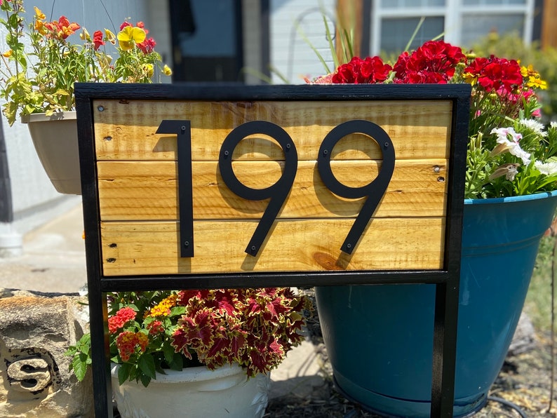 May include: Black metal house number sign with the number 199 on a wooden background. The sign is mounted on a black metal frame and is surrounded by flowers.