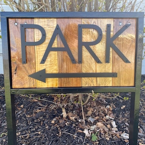 May include: A wooden sign with the word "PARK" in black metal letters and an arrow pointing left. The sign is made of vertical wooden planks with varying shades of brown and gold, framed by a black metal border and supported by a black metal post.