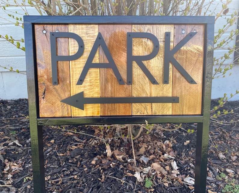 May include: A wooden sign with the word "PARK" in black metal letters and an arrow pointing left. The sign is made of vertical wooden planks with varying shades of brown and gold, framed by a black metal border and supported by a black metal post.