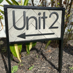 May include: A black metal sign with the text "Unit 2" and an arrow pointing left. The sign has a gray, textured background with a black border and is mounted on a black metal post. The sign is outdoors.