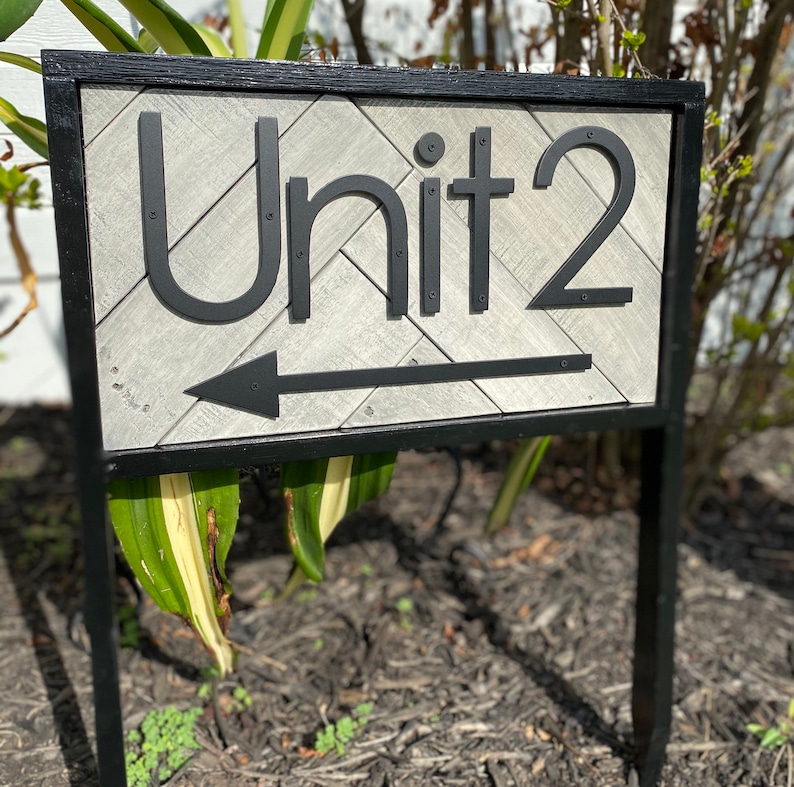 May include: A black metal sign with the text "Unit 2" and an arrow pointing left. The sign has a gray, textured background with a black border and is mounted on a black metal post. The sign is outdoors.