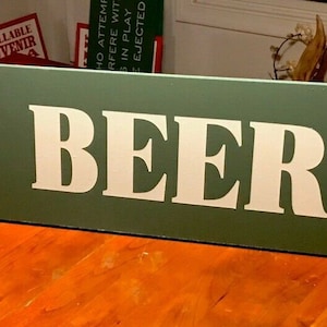 May include: A green wooden sign with white lettering that reads "COLD BEER" and a white pointing hand.