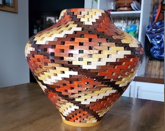 Puede incluir: Un jarrón de madera con un patrón geométrico en tonos marrones, naranjas y amarillos. El jarrón está hecho de múltiples piezas de madera que están entrelazadas de forma intrincada.
