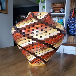 May include: A wooden vase with a geometric pattern in shades of brown, orange, and yellow. The vase is made of multiple pieces of wood that are intricately woven together.