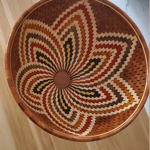 May include: A wooden bowl with a geometric pattern in shades of brown, orange, and yellow. The bowl is made of multiple pieces of wood that are intricately woven together to create a starburst design.