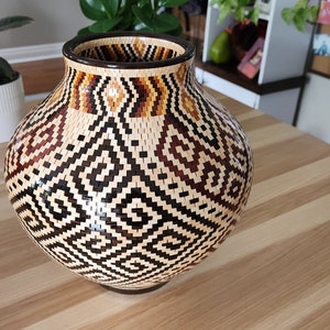Puede incluir: Un jarrón grande y redondo con un patrón geométrico en tonos marrones, negros y beige. El jarrón está hecho de madera y tiene un acabado liso y pulido.