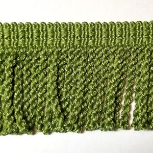 Puede incluir: Primer plano de un ribete de flecos verdes. El ribete presenta una sección superior tejida con una serie de bucles verticales y una franja retorcida texturizada que cuelga por debajo. El color es un verde medio vibrante.