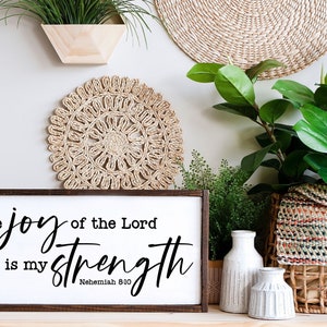 May include: A wooden sign with a white background and black text that reads "the joy of the Lord is my strength" with the scripture reference "Nehemiah 8:10" below. The sign is sitting on a table with a plant and two white vases.