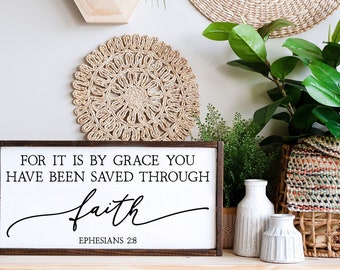 May include: A wooden sign with a brown frame and white background. The sign has black text that reads "FOR IT IS BY GRACE YOU HAVE BEEN SAVED THROUGH faith EPHESIANS 2:8".