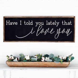 May include: A black framed sign with white text that reads "Have I told you lately that I love you" hanging on a white wall above a wooden tray filled with green eucalyptus and white cotton stems.