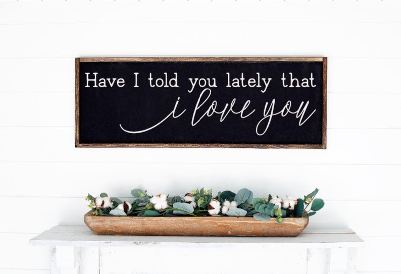 May include: A black framed sign with white text that reads "Have I told you lately that I love you" hanging on a white wall above a wooden tray filled with green eucalyptus and white cotton stems.