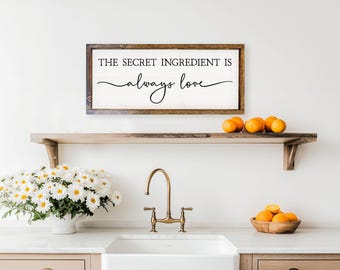 May include: A kitchen scene featuring a wooden shelf with oranges and a framed sign that reads "THE SECRET INGREDIENT IS always love." A white sink, faucet, and a vase of white flowers complete the decor.