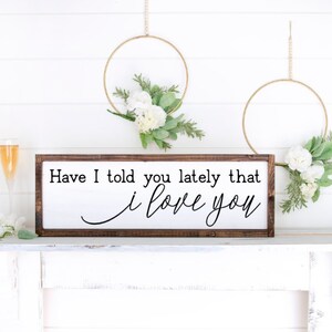 May include: A white wooden sign with a brown frame, hanging on a white mantel. The sign reads "Have I told you lately that I love you".