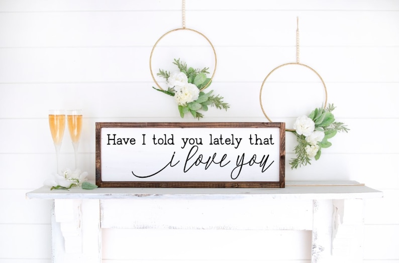 May include: A white wooden sign with a brown frame, hanging on a white mantel. The sign reads "Have I told you lately that I love you".