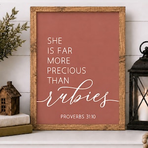 May include: A rectangular wall art print in a rustic wooden frame featuring a terracotta background with white text that reads: SHE IS FAR MORE PRECIOUS THAN rubies PROVERBS 31:10. The print is displayed upright on a flat surface.