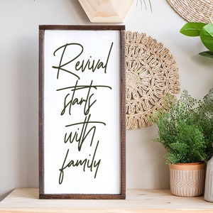 May include: A wooden sign with a brown frame and white background. The sign has black calligraphy that reads "Revival starts with family".