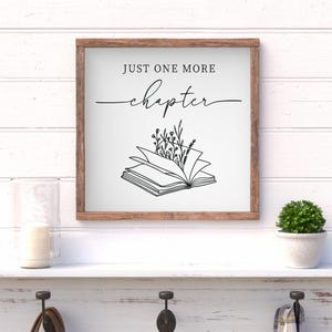 May include: Wooden framed sign with the text "JUST ONE MORE chapter" above an illustration of an open book with floral accents. The sign is displayed on a white shelf with a small potted plant and a glass.