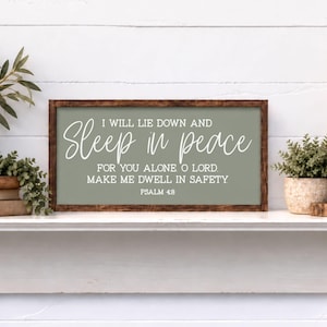May include: A rectangular wooden sign with a dark brown frame and a sage green background. The text in white reads: I WILL LIE DOWN AND Sleep in peace FOR YOU ALONE O LORD. MAKE ME DWELL IN SAFETY PSALM 4:8. The sign is displayed on a white mantel.