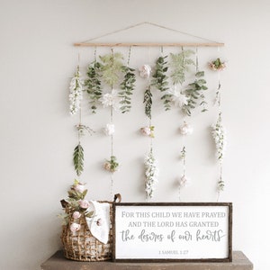 May include: A wooden sign with a white background and gray text that reads "FOR THIS CHILD WE HAVE PRAYED AND THE LORD HAS GRANTED THE DESIRES OF OUR HEARTS 1 SAMUEL 1:27". The sign is hanging on a wall with a floral garland behind it.