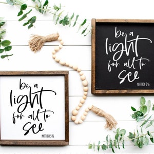 May include: Two square wooden signs with the text "be a light for all to see" in white script. One sign has a black background, the other is white. Both have a brown wooden frame. Decorative elements include eucalyptus and wooden beads.