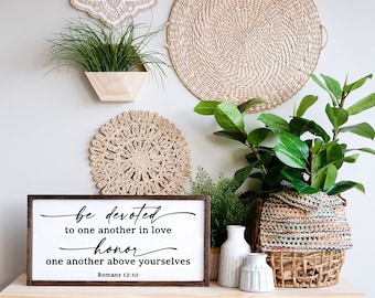May include: A wooden sign with the text "be devoted to one another in love honor one another above yourselves Romans 12:10" in black script font. The sign is on a wooden table with two white vases and a woven basket with a green plant.