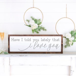 May include: A wooden sign with the text "Have I told you lately that I love you" in gray lettering. The sign is framed in dark brown wood and is sitting on a white shelf. There are two gold hoops with white flowers hanging on the wall behind the sign.