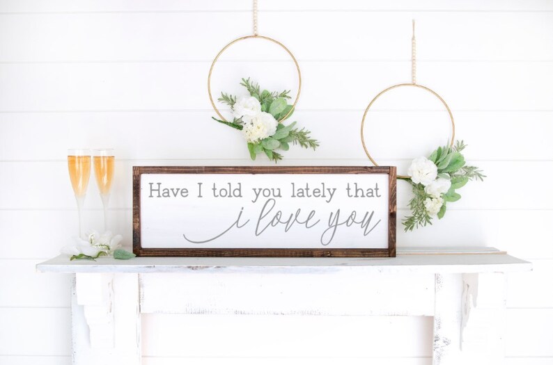 May include: A wooden sign with the text "Have I told you lately that I love you" in gray lettering. The sign is framed in dark brown wood and is sitting on a white shelf. There are two gold hoops with white flowers hanging on the wall behind the sign.