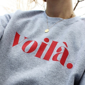 Könnte beinhalten: Ein graues Sweatshirt mit dem Wort "Voilà" in roter, stilisierter Schrift. Das Sweatshirt ist aus einem weichen Material gefertigt und hat einen Rundhalsausschnitt. Eine goldene Halskette mit Anhänger ist sichtbar.