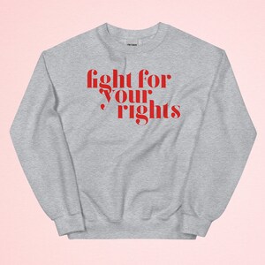 Könnte beinhalten: Ein hellgraues Sweatshirt mit dem roten Schriftzug "fight for your rights" in einer auffälligen, stilisierten Schriftart. Das langärmlige Oberteil ist aus weichem Material und für den Freizeitgebrauch geeignet.