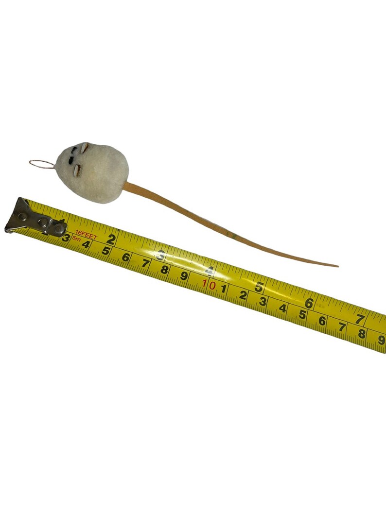 Pode incluir: Um brinquedo de rato de feltro branco com uma cauda de madeira marrom, medido com uma fita m&eacute;trica amarela. O rato mede aproximadamente 25 cm de comprimento.