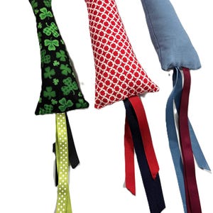 May include: Three cat toys with different fabric patterns and colors. Each toy has a long tail with multiple ribbons. The toys are green with shamrocks, red with white circles, and blue.