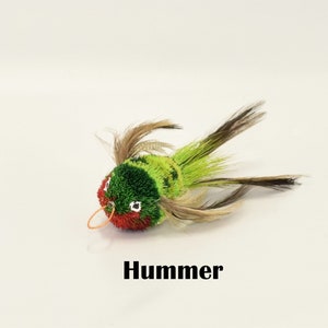 May include: A green, red, and brown feathered toy bird with a black elastic loop. The toy is labeled "Hummer".
